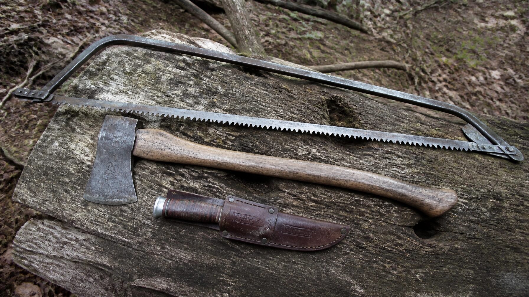 bushcraft gear photo showing a well-worn cutting kit on a rough log in the woods: an old refurbished axe with honest patina, a practical metal bow saw, and an Old Hickory knife in a worn leather sheath. Natural daylight, true-to-life colors, and a plain forest background